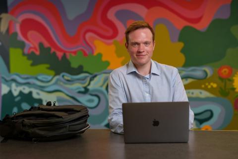 Kieran sits facing the camera with his laptop open and bookbag nearby on the table. He smiles in a light blue shirt against a colorful mural backdrop depicting a river, trees, and a sunset with swirly abstract lines. 