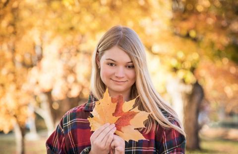Courtney, 一个长着金色头发的女人, 身穿红色法兰绒衬衫站在秋叶丛中，手持叶扇对着镜头